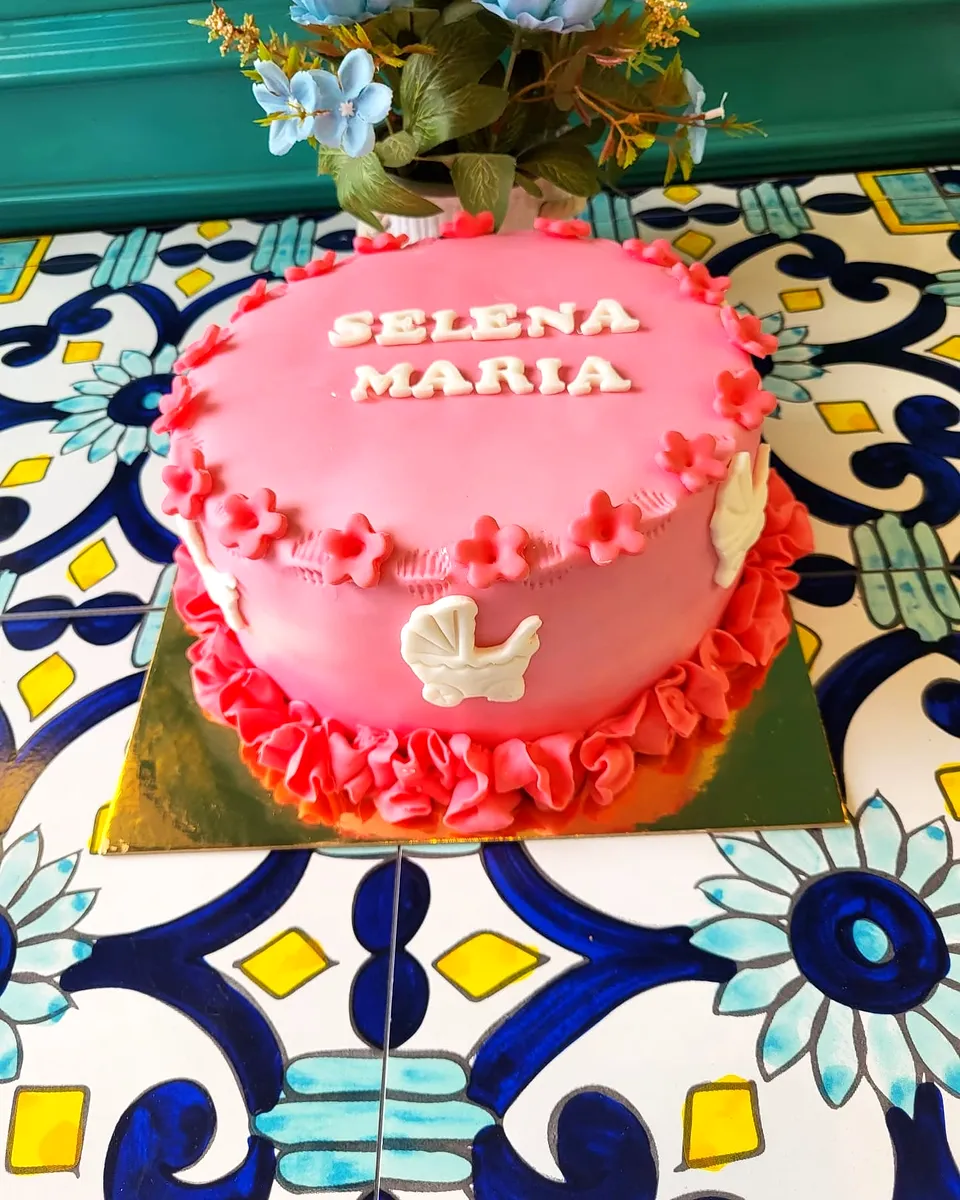 Tort de botez roz cu volănașe, cu numele Selena Maria scris pe blat și un cărucior alb din fondant