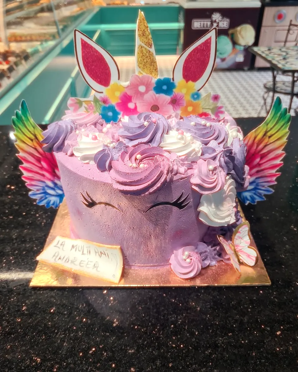 Tort unicorn mov, cu aripi colorate, corn auriu și flori din cremă